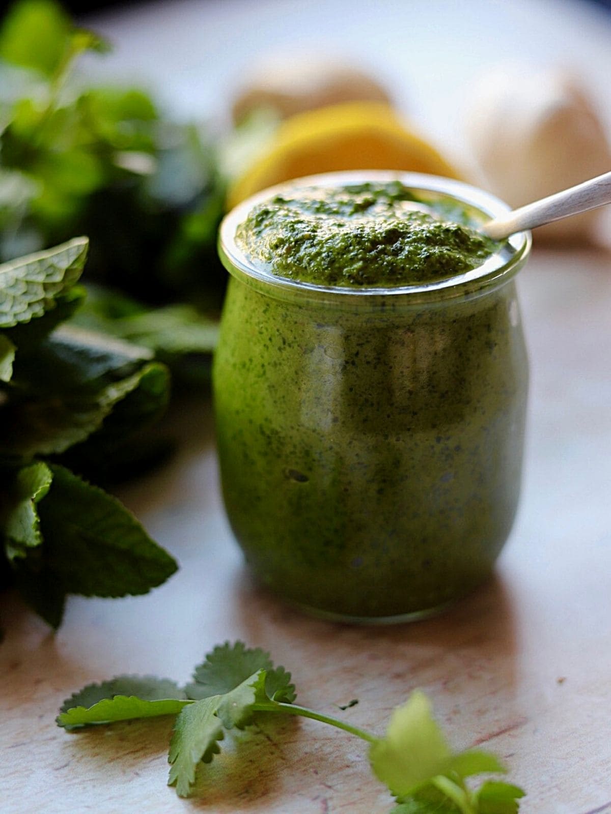 Green chutney in glass jar with spoon surrounded by fresh mint and lemon.