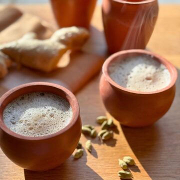 Two earthenware cups of masala chai next to cardamom pods and a ginger root.