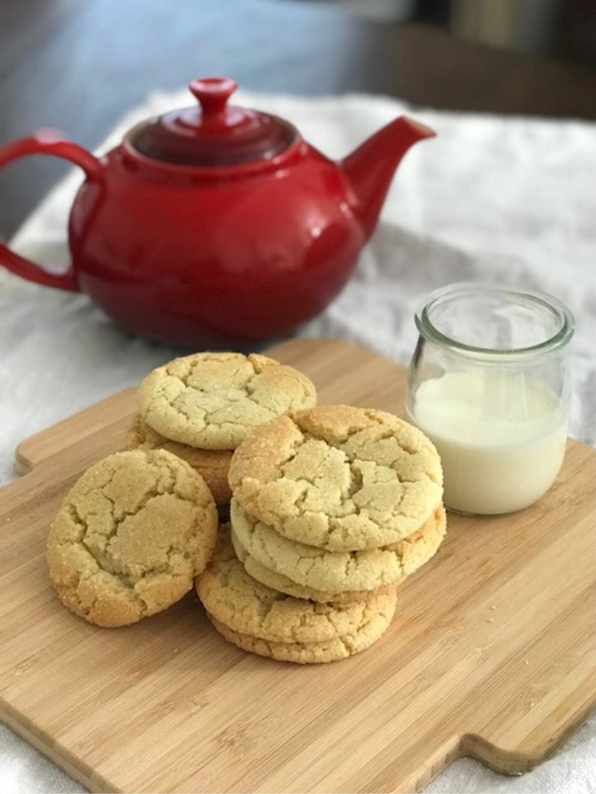 A pile of sugar cookies on a wooden board with a red teapot and beaker of milk.