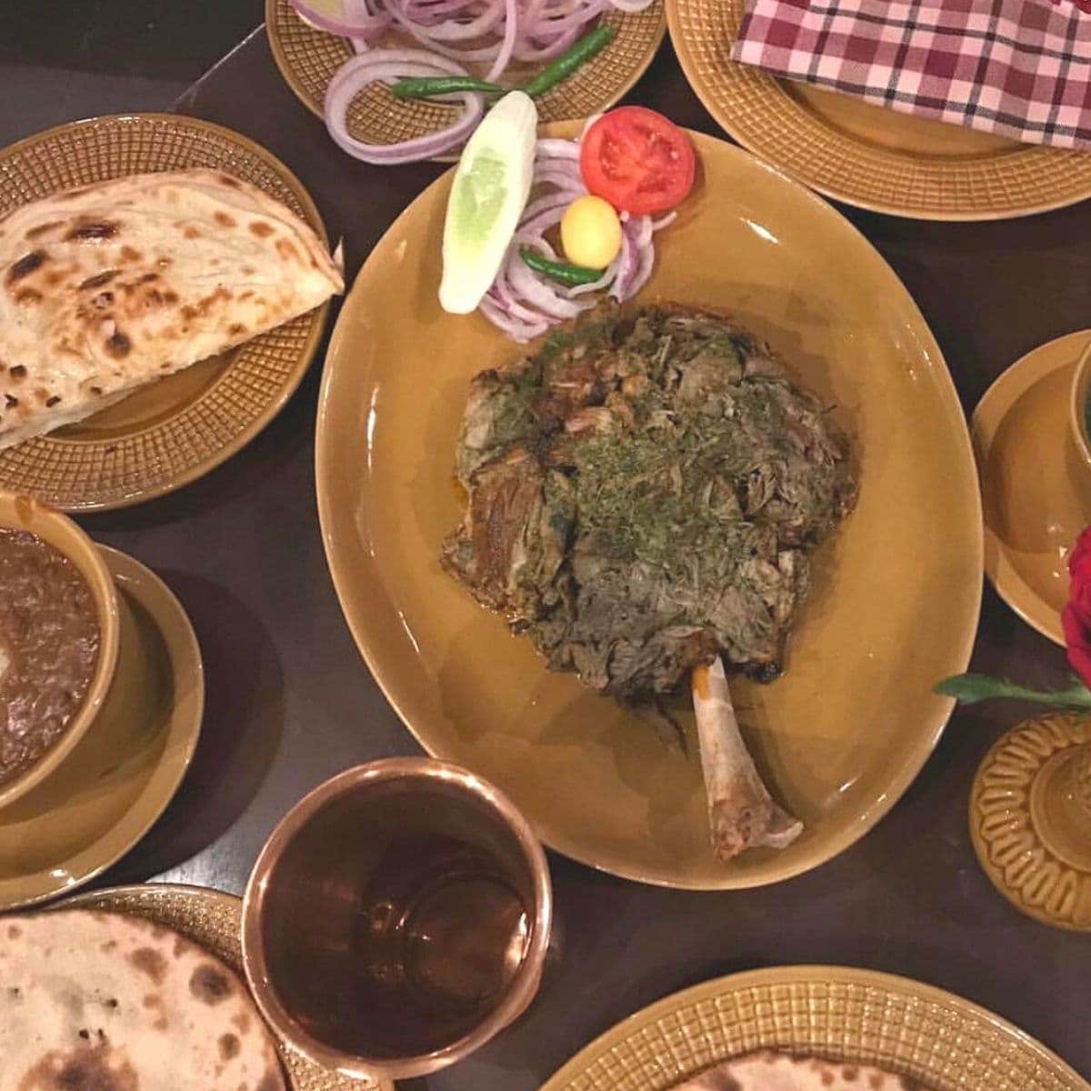 Leg of goat meat on platter on a dining table surrounded by plates and dishes.