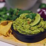 Image of guacamole in small black bowl garnished with cilantro and slice of lime.