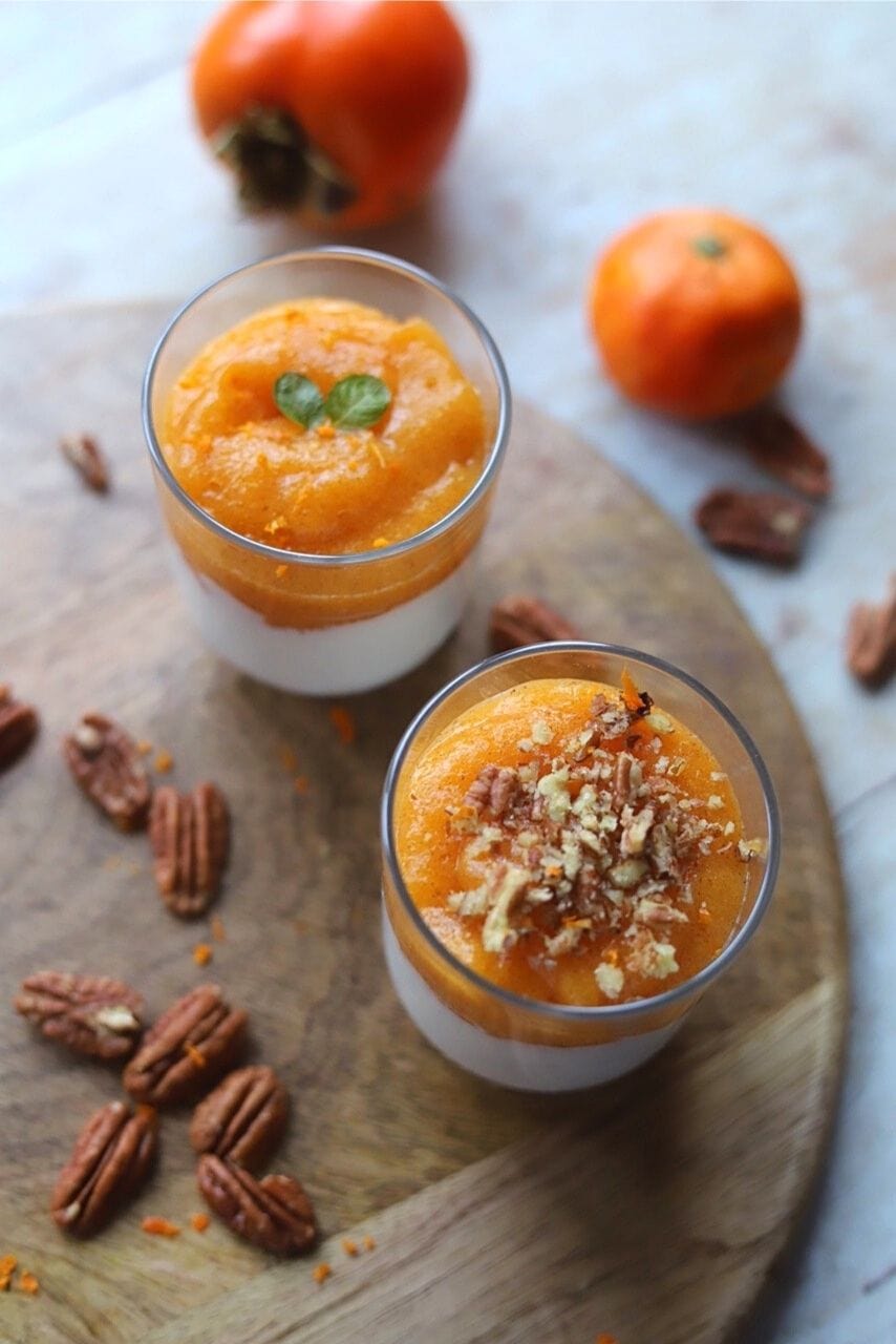 Persimmon and ricotta dessert in two glasses surrounded by pecans and full persimmons.