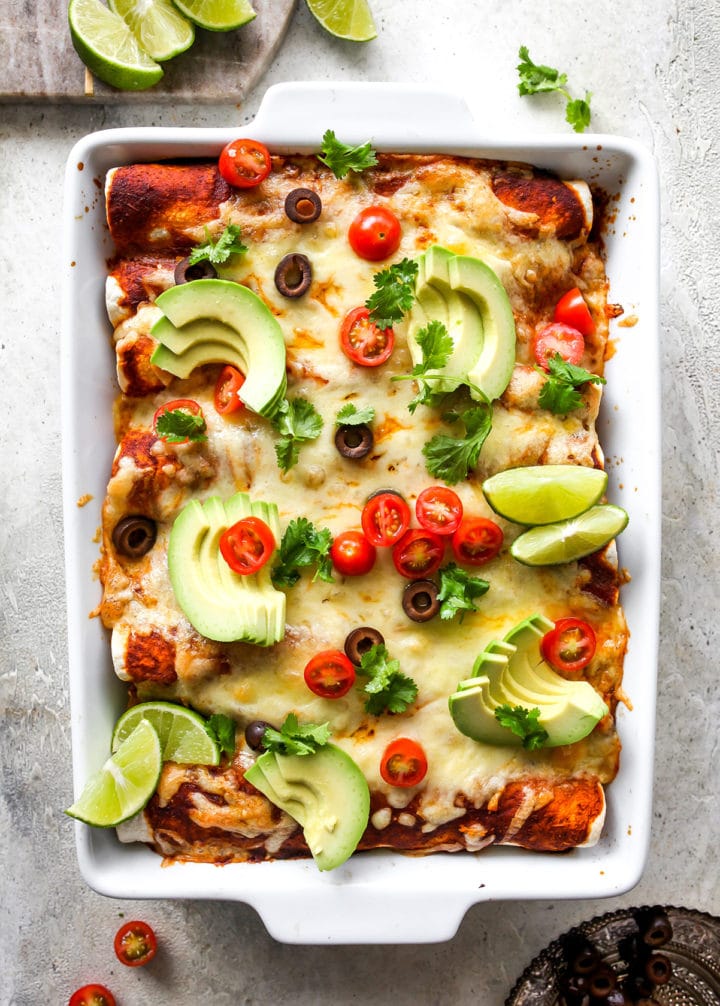 Image of baked enchiladas with melted cheese and sliced raw avocado in white casserole dish.