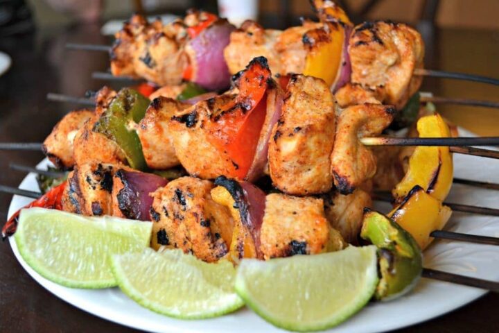 Image of skewers of chicken and vegetable kebabs piled high on plate and lime wedges.