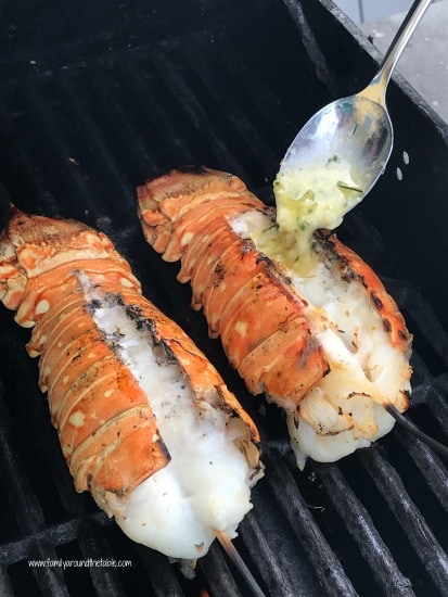 Image of two lobster tails on the grill being dressed with garlic butter.