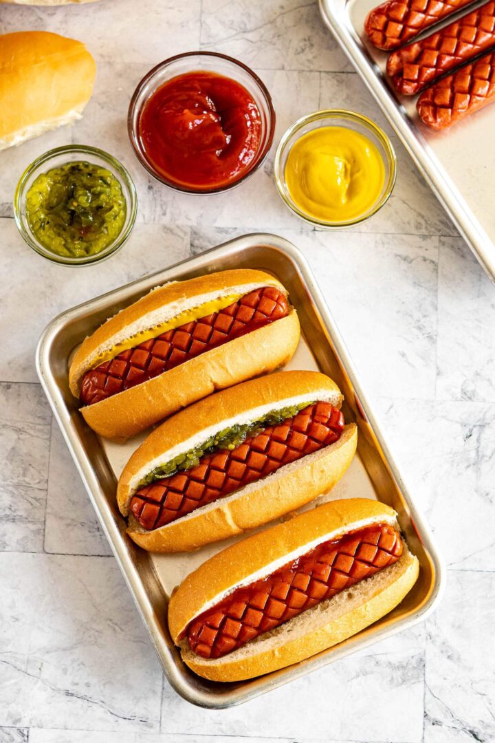 Image of three smoked hot dogs with diamond cuts in buns filled with different sauces.