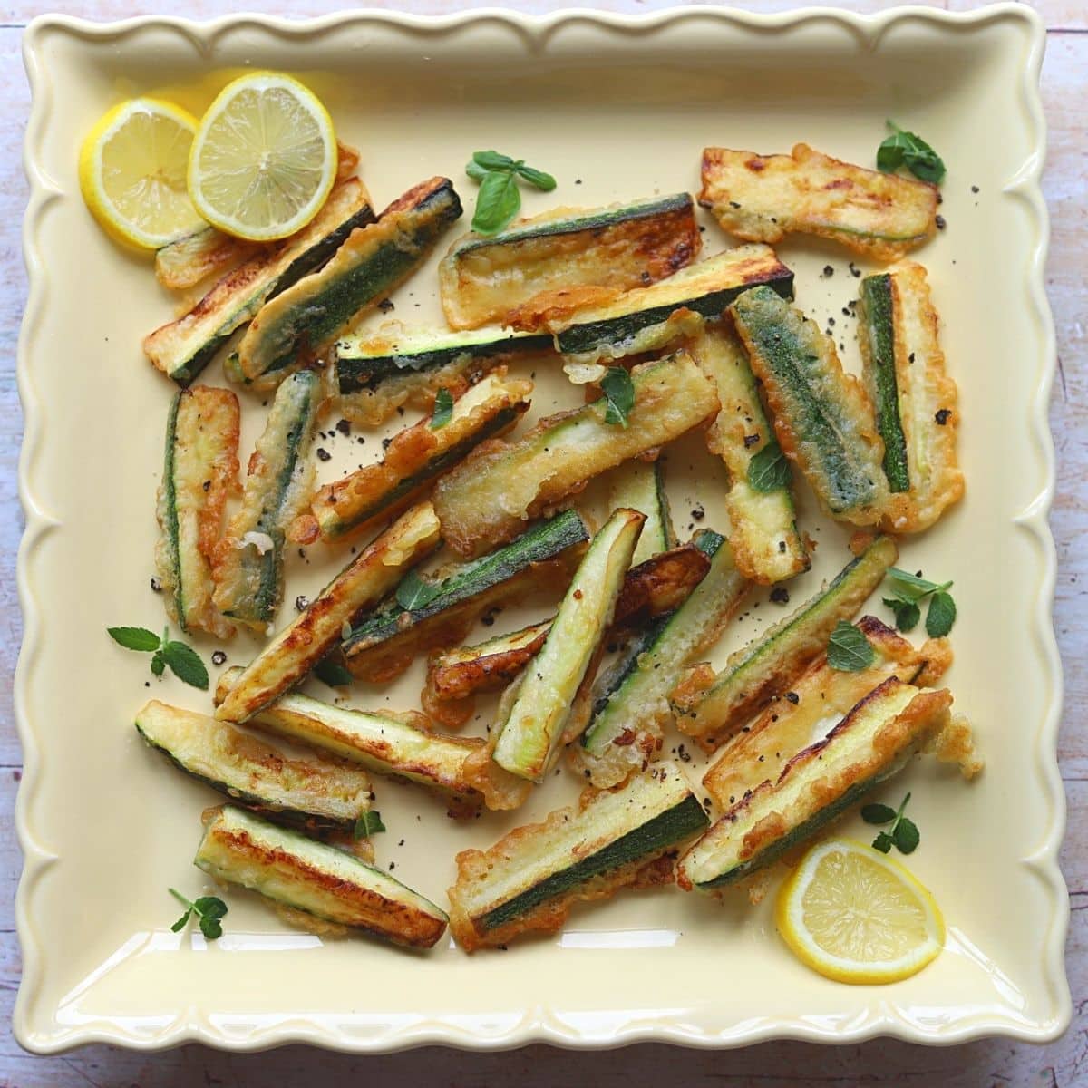 Fried zucchini chips on a large yellow platter with sliced lemon and fresh mint.