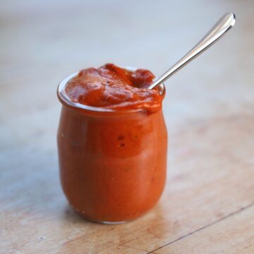 Red pepper dip in glass jar with metal spoon.