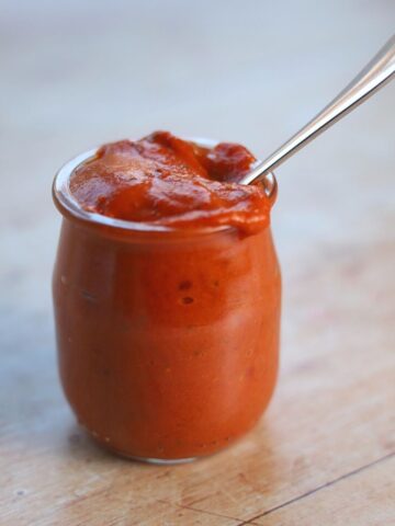 Red pepper dip in glass jar with metal spoon.