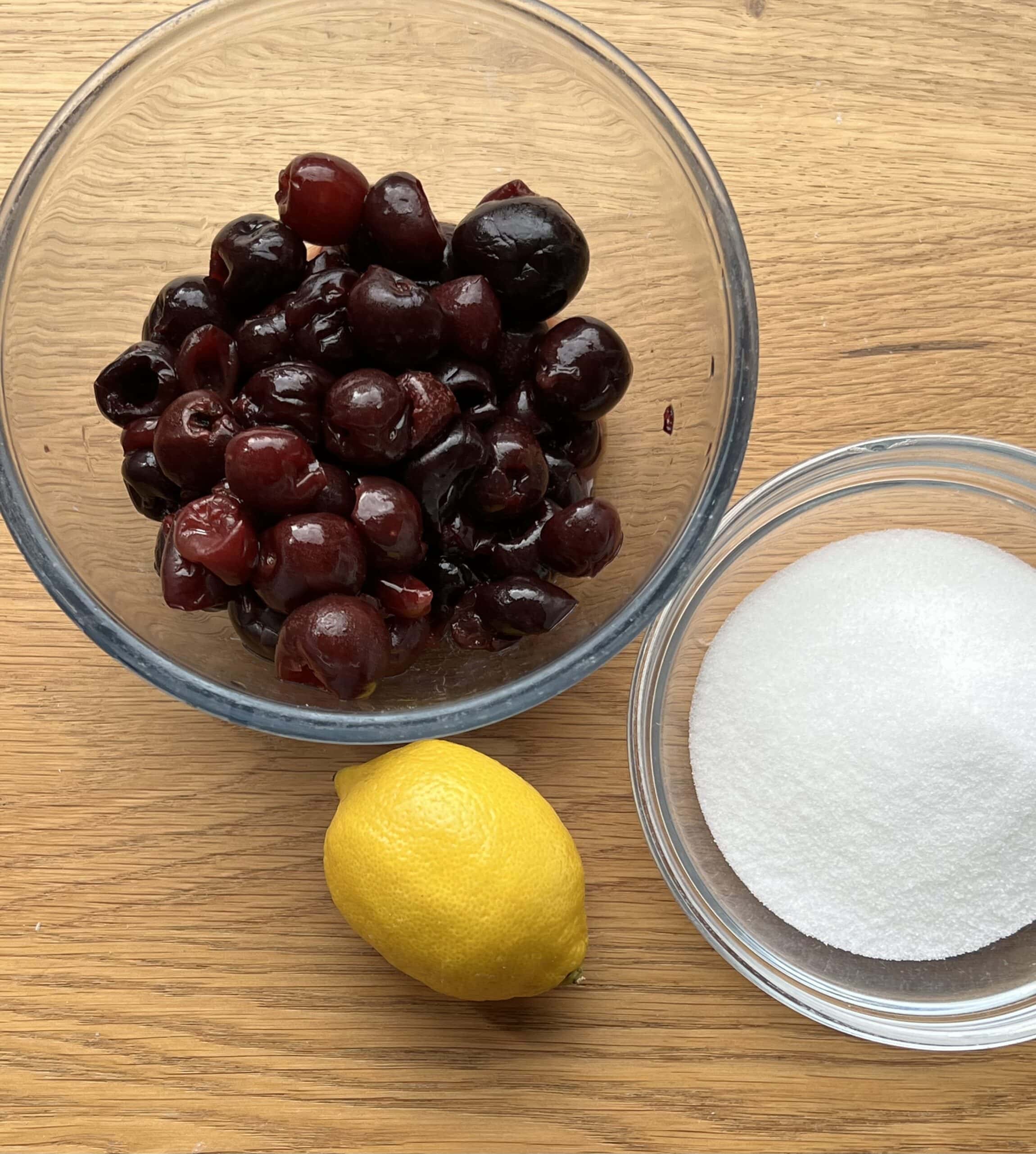 Bowls of cherries, sugar and a lemon on wooden counter.