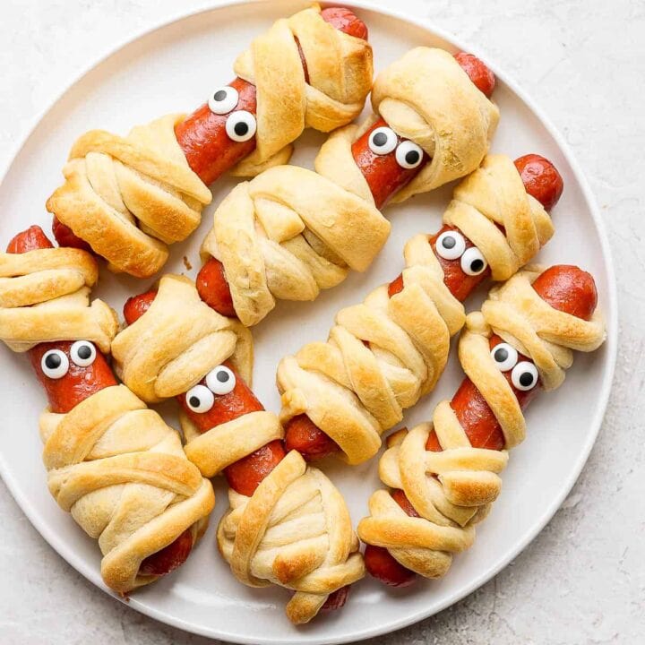Six hot dogs wrapped in baked pastry with candy eyes on white plate.