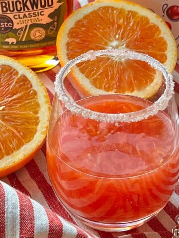 Orange drink next to halved oranges on red stripe cloth.