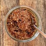 Chicken rub in small glass jar with spoon on wooden board.