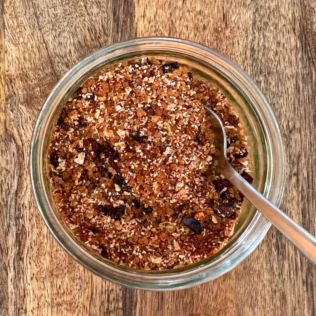 Chicken rub in small glass jar with spoon on wooden board.